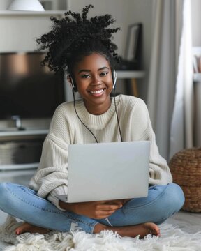 Beautiful young African American girl with headphones and laptop sitting with folded legs on the floor in a cozy room. Smiling black female student learning online. Remote education concept.