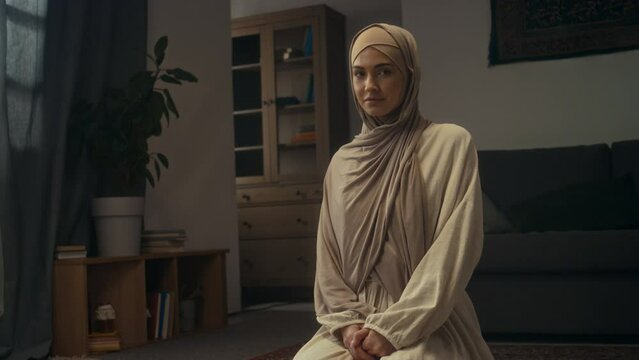 Medium portrait of attractive young muslim woman in light beige hijab and abaya posing sitting on knees on floor in majlis and looking at camera