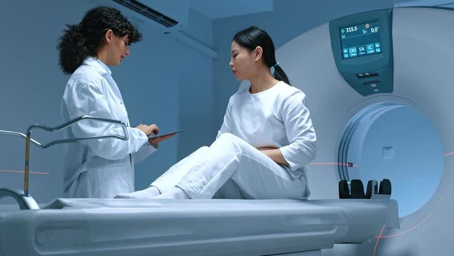 Examination of magnetic resonance imaging at background of modern equipment. Patient sitting at computerized tomography scanner bed, complaining at whole body pain, listening to doctor attentively.
