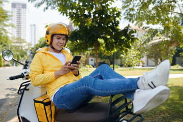 Smiling courier sitting on scooter, resting between rides, texting friends