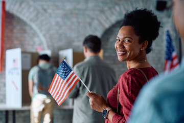 Happy black woman with US flag waiting to vote during elections and looking at camera.