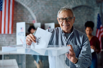 Happy senior man inserting his ballot into box while voting during US elections and looking at...