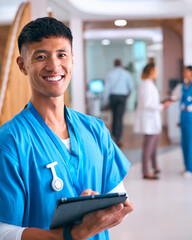 Portrait Of Smiling Male Doctor Or Nurse Wearing Scrubs In Hospital With Colleague In Background