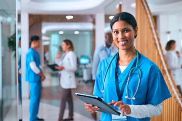 Female Doctor Or Nurse In Scrubs With Digital Tablet In Hospital With Colleagues In Background