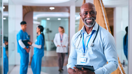 Portrait Of Mature Male Doctor With Digital Tablet In Hospital With Colleagues In Background - 750766300