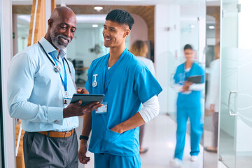 Male Doctor And Nurse In Scrubs With Digital Tablet Meeting In Hospital With Colleagues Behind