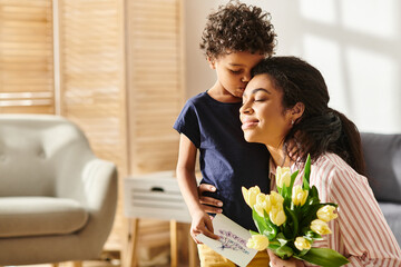 joyous african american woman hugging her son and holding postcard and tulips, modern parenting