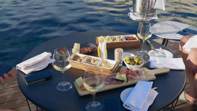 Friends having aperitif time on the swimming pool side, people sitting at the table and tasting delicious traditional Italian aperitif, goblets with refreshing white wine, salami and cheese slices