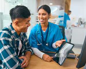 Female Doctor Wearing Scrubs At Hospital Appointment With Male Patient Looking At Digital Tablet
