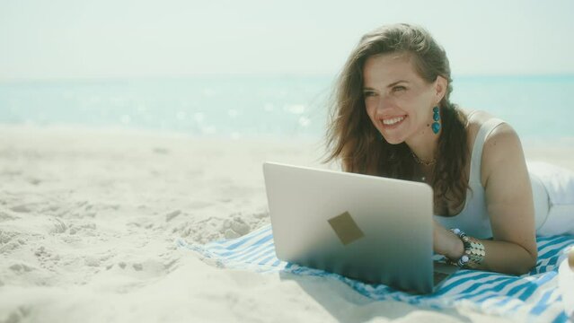 smiling modern 40 years old woman on the ocean coast with straw bag and striped towel using laptop.