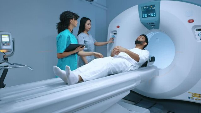 Diverse group of female neurologist neobiologist with male patient in room of computed tomography. Medical worker putting buttons on MRI, woman doctor standing near patient and processing procedure.