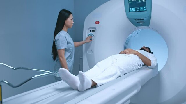 Pretty female doctor performs CT scan for male patient at diagnostic clinic. Medical girl nurse putting buttons on MRI device. Magnetic resonance imaging in study of human body. Medicine, healthcare.