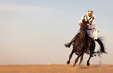 Saudi man in teaditional clothing riding a black horse in a desert