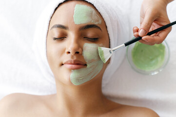 Facial treatments, wrinkle prevention, facial cleansing. Top view of beauty procedure, therapist applying green face mask on the face of a beautiful brazilian or hispanic young woman