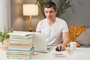 Focused concentrated Caucasian adult man freelancer wearing white T-shirt working at home with laptop typing on keyboard looking at monitor doing his online project