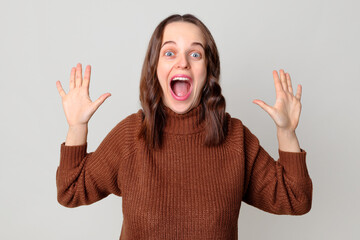 Extremely happy cheerful Caucasian brown haired woman wearing brown jumper standing isolated over light gray background raising her hands showing palms rejoicing her success