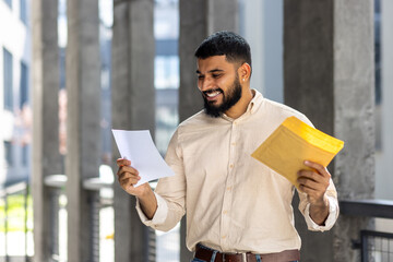 Happy young Indian man standing outside building, holding envelope and smilingly reading received...
