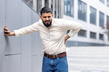 A tired Muslim man is standing outside leaning against the wall of the building, holding his hand behind his back, grimacing and feeling severe pain in his lower back - obrazy, fototapety, plakaty