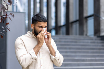 Close-up photo of a young Indian man standing near a building on the street and wiping his nose with a tissue, suffering from a severe runny nose and seasonal allergies
