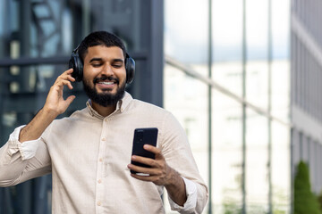 A young Indian man is standing on the street in a shirt and headphones, holding a phone in his...