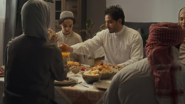 Rear footage of big islamic family having festive Eid dinner together at home. Young man pouring orange juice into glasses and serving relatives