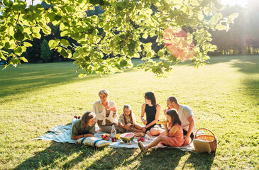 Big family under Linden tree on the picnic blanket on the in city park green grass. They are eating...