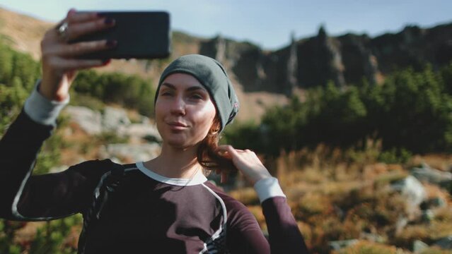 Active tourist woman holding cellphone to take selfie picture. Mountain nature landscape in background. Caucasian girl hiker enjoys moment. Travel, tourism, holiday, trekking, hiking, active lifestyle