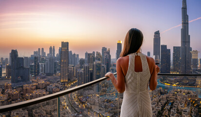 A beautiful luxury woman in a white dress enjoys the sunset view behind the modern skyline of Downtown Dubai, UAE, with a drink