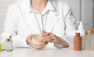 Dermatologist with jar testing cosmetic product at white table indoors, selective focus