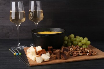 Fondue pot with melted cheese, glasses of wine and different products on black wooden table