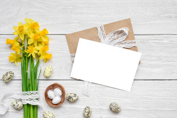 Easter greeting card with spring yellow Narcissus flowers and easter colorful eggs on white wooden background. Flat lay. Mock up. Top view with copy space