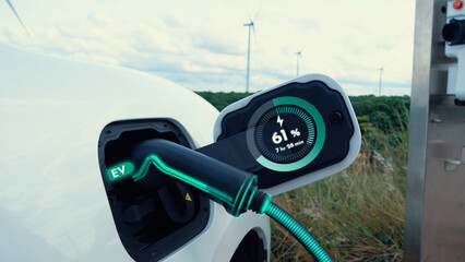 Electric car recharging energy from EV charging station display futuristic smart battery status hologram by EV charger plug cable in wind turbine farm. Alternative clean energy sustainability. Peruse