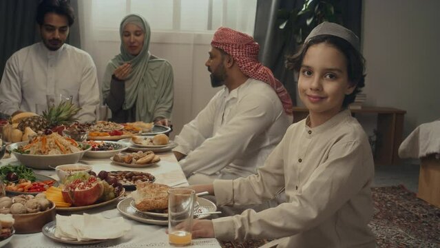 Side slowmo portrait of little Biracial boy looking at camera while sitting at festive table with relatives on Uraza Bayram celebration at home