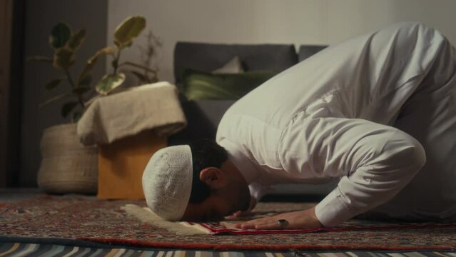 Side ground level footage of muslim man in cap and thawb prostrating while praying to Allah on prayer map at home