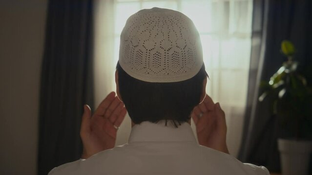 Rear chest up footage of anonymous muslim man in white skullcap raising hands and reciting salat while praying takbirs at home