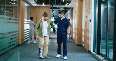 Caucasian kind physiotherapist supporting and talking with man using crutches. Medical workers spending time communicating and assisting people in contemporary hospital.