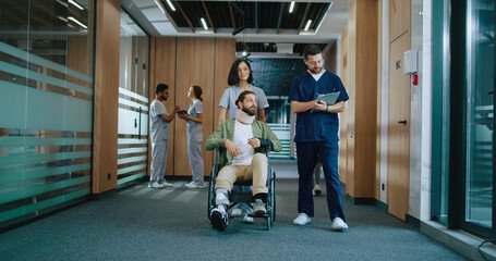 Young beautiful nurse and handsome professional doctor in corridor helping injured Caucasian patient in wheelchair consulting and providing healthcare. Concept of hospital.