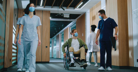 Injured Caucasian male patient in wheelchair moving around hospital corridors with professional helpful doctor in uniform holding x-ray talking and consulting man. Modern clinic concept.