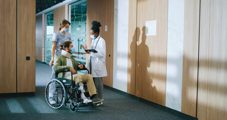 Young Caucasian bearded man sitting in wheelchair and talking with helpful medical staff in modern...