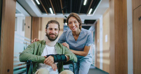 Portrait of happy injured Caucasian man recovering in hospital with qualified female doctor looking at camera and smiling. Bearded young patient and helpful nurse in modern clinic.