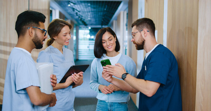 Side view portrait of diverse group of young skilled medical professionals chatting in modern hospital corridor during break. In uniform successful medical workers conversing and having fun.