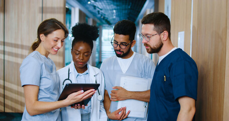 Multicultural group of youthful medical personnel communicating in modern hospital hallway during...