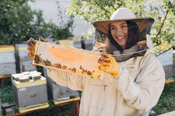 Happy female Beekeeper in uniform standing in apiary and holding honeybee frame
