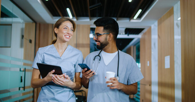 Portrait of skilled happy doctors walking in modern hospital hallways smiling and talking about their jobs. Caucasian doctor and young cheerful male Indian nurse having coffee.