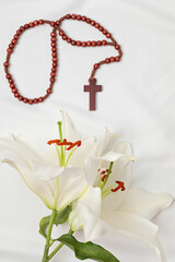 A symbolic Christian composition with christian cross lies beside a fresh white lily on a white fabric background as traditional Easter symbols.