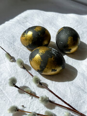 Dark blue and gold Easter eggs lie on a white light tablecloth under the rays of the sun, willow branches lie nearby. An Easter card. Vertical photo