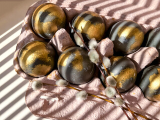 Stripes of light and shadow from the blinds lie on the black and gold Easter eggs that are in the tray, next to the willow branches. Preparing for the Easter holiday