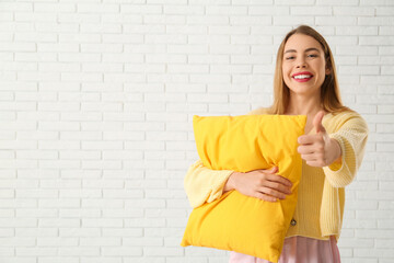 Obraz premium Beautiful young woman holding yellow soft pillow and showing thumb-up gesture on white brick background