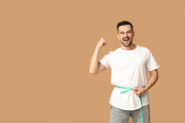 Handsome sporty young man measuring his waist on brown background. Weight loss concept - obrazy, fototapety, plakaty