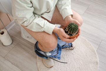Young man with cactus sitting on toilet bowl in restroom, closeup. Hemorrhoids concept - obrazy, fototapety, plakaty